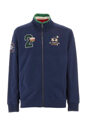 Giacca di felpa con zip color navy realizzata in tessuto morbido; presenta rifiniture al collo verde e rosso, loghi ricamati e tasche frontali. La forma è attillata con polsini a coste.