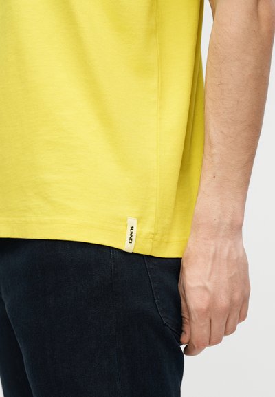 Ανδρικός βραχίονας δίπλα στο τελείωμα ενός κίτρινου T-shirt με μικρή λευκή ετικέτα, σε συνδυασμό με σκούρο μπλε παντελόνι, πάνω σε απλό φόντο.