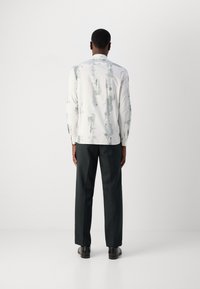 Λευκό πουκάμισο με ανοιχτό πράσινο αφηρημένο σχέδιο, μακριά μανίκια και άνετη εφαρμογή, συνδυασμένο με σκούρες, ραμμένες παντελόνες και μαύρα παπούτσια.