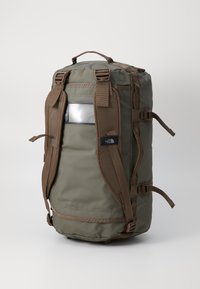 Olivengrønn duffel ryggsekk med brune stropper, topphåndtak og en klar lomme. Inkluderer sidetilpassere og slitesterkt stoff for styrke.