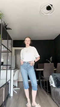 Fehér grafikus póló, kék farmer cropped nadrág és bézs szandál. Laza haj, áll egy modern belső térben növényekkel és bútordarabokkal.
