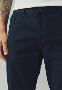 Tamno plave pamučne hlače uskog kroja. Imaju petlje za pojas, prednju džep i vidljivo kopčanje gumba blizu struka.