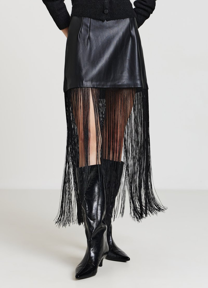 Kurzer schwarzer Lederrock mit langen schwarzen Fransen, kombiniert mit schwarzen, spitz zulaufenden High-Heel-Stiefeln an einem Model.