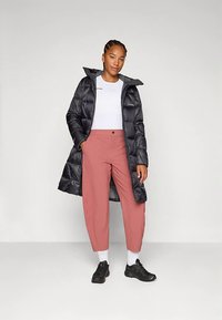 Mulher vestindo um casaco preto longo tipo puffer, uma t-shirt branca, calças cor-de-rosa, meias brancas e sapatilhas pretas, de pé com as mãos nos bolsos.