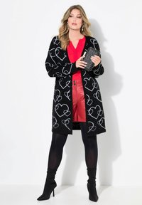 Schwarze Strickjacke mit weißen Herzmustern, rotes Oberteil und rote Leder-Shorts. Schwarze High-Heel-Stiefel vervollständigen das Outfit, dazu eine strukturierte Clutch-Handtasche.