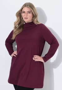 Burgundské dlouhé šaty s rolákem vyrobené z měkké tkaniny, s uvolněným střihem a kulatým lemováním. Modelka nosí oversized brýle.