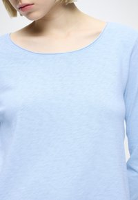 Jasnoniebieska koszula z długim rękawem, z szerokim, zaokrąglonym dekoltem i miękką, fakturowaną tkaniną, widoczna od ramion w dół.