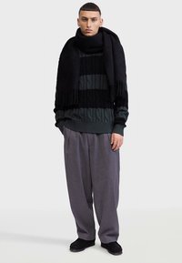 Pull en maille rayé noir et vert foncé avec des motifs torsadés, associé à un pantalon gris ample et une longue écharpe noire à franges.