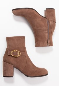 Bottes chelsea en suede brun avec un talon épais, fermeture éclair sur le côté et un accent en boucle dorée sur la sangle. Texture lisse et design à bout rond.