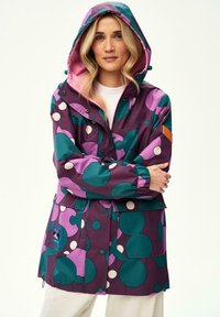 Kobieta o blond włosach, ubrana w fioletową kurtkę z kapturem, ozdobioną dużymi zielono-białymi groszkami, stoi z założonymi rękami.