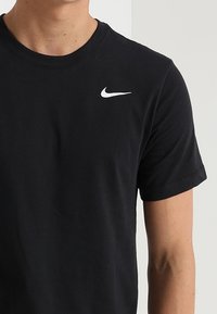 Muž v jednoduchém černém tričku Nike s bílým logem Nike swoosh na levé horní části hrudi.