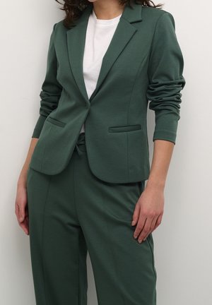 Femme portant un blazer vert forêt et un pantalon assorti sur un t-shirt blanc, sur un fond uni.