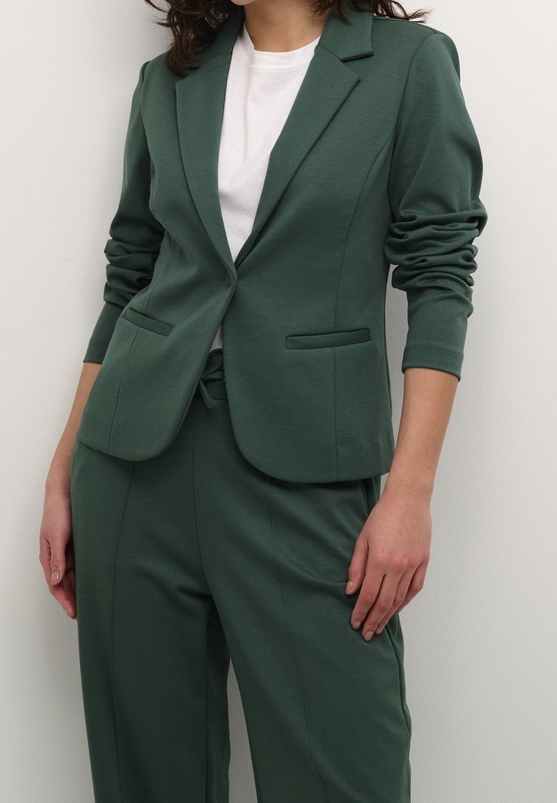 Femme portant un blazer vert forêt et un pantalon assorti sur un t-shirt blanc, sur un fond uni.