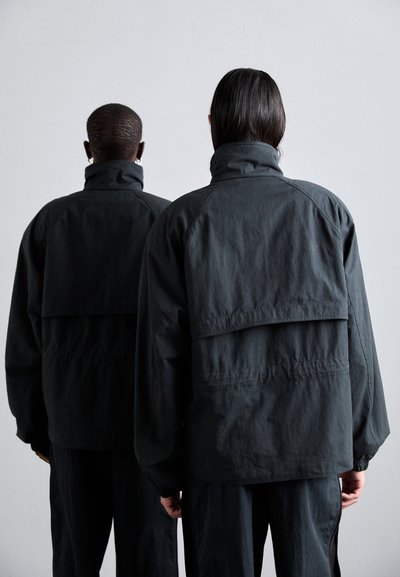 Black oversize jackets with high collars, featuring a layered back design and textured fabric, worn by two models from behind.