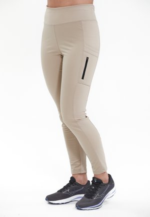 Beiga leggings med hög midja och sidoficka med dragkedja, tillverkade av ett mjukt och stretchigt material. Kombineras med svarta träningsskor.