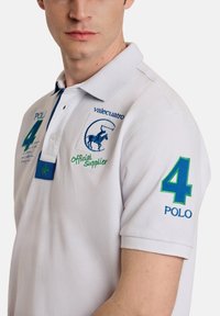 Camisa polo blanca con cuello, que presenta bordados en azul y verde. Los detalles notables incluyen el número "4" y el nombre de la marca "valecuatro."