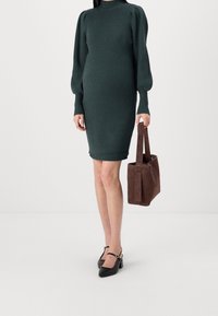 Robe en tricot vert foncé avec une texture côtelée, un col haut et des manches longues bouffantes. Associée à un sac fourre-tout en daim marron et des chaussures noires.