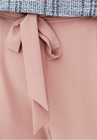 Un tissu rose pâle avec une texture lisse présente un détail de nœud noué à la taille, complété par un bord supérieur à motifs multicolores.
