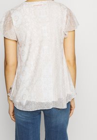 Blouse beige clair et transparente avec manches volantes, présentant un motif subtil et un ourlet légèrement évasé. Logo de la marque au dos du col.
