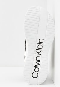 Černé tenisky s texturovanou bílou gumovou podrážkou, s logem "Calvin Klein" v černé barvě na patě, a elegantním designem s minimem akcentů.