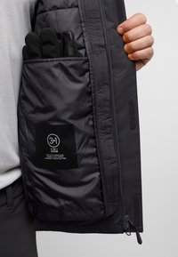 Zwarte geïsoleerde jas met een ritsvak voor handschoenen. Inclusief een label dat een 3-in-1 systeem voor bescherming tegen het weer aangeeft.