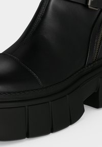 Gros plan d'une botte en cuir noir avec une semelle épaisse et volumineuse et une fermeture éclair latérale, montrant des coutures détaillées et la texture.