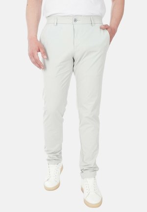 Uomo che indossa pantaloni aderenti grigio chiaro, scarpe da ginnastica bianche e una camicia bianca, con una mano in tasca e in piedi su uno sfondo bianco.