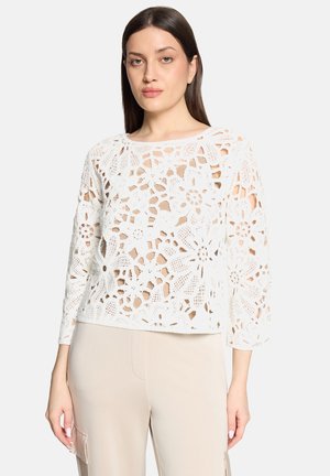 Femme portant un haut long à manches longues en crochet floral blanc et un pantalon beige, debout devant un fond clair uni.