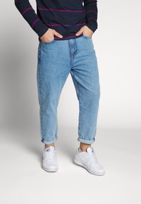 Ljust blå denimjeans med en avslappnad passform och uppvikta muddar, kombinerade med vita sneakers och en marinblå långärmad skjorta med röda ränder.