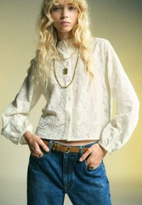 Blusa bianca, texturizzata, con maniche lunghe e gonfiate, chiusura con bottoni. Abbinata a jeans blu a vita alta e cintura marrone. Collane d'oro sovrapposte.