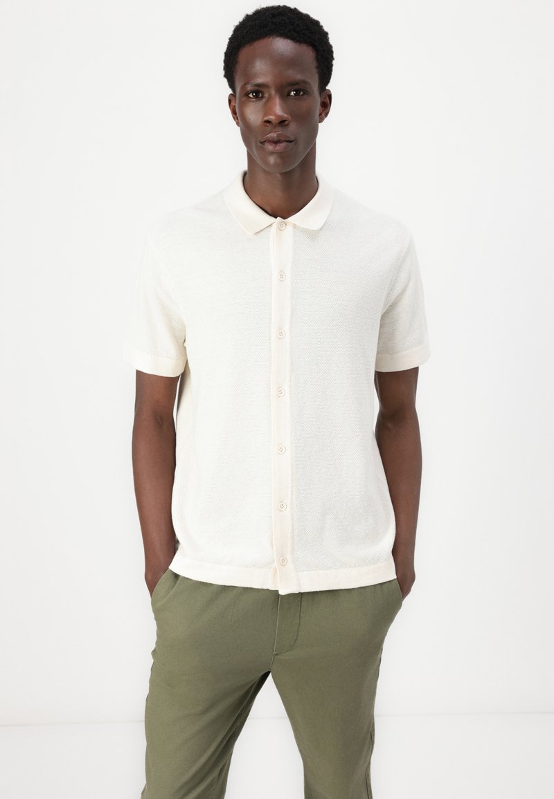 Polo de punto de manga corta en color beige claro, con cuello de solapa, cierre de botones en la parte delantera y un cuello en un tono pálido contrastante, combinado con pantalones verde oliva.