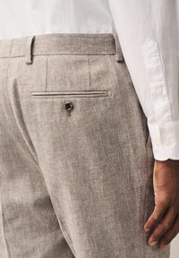 Pantalones de lino beige con una superficie texturizada, que cuentan con un bolsillo trasero con cierre de botón y costuras en contraste. Manga de camisa blanca visible.