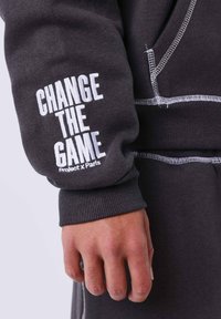 Sudadera gris con el texto blanco "CAMBIA EL JUEGO" y "Project x Paris" en la manga. Presenta costuras en contraste y puños acanalados.