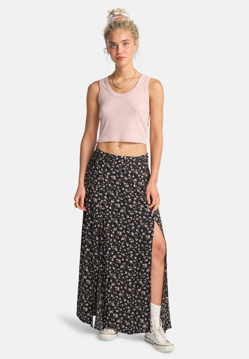Helles rosa, kurzes Tanktop kombiniert mit einem schwarzen, floralen Maxikleid mit seitlichen Schlitzen. Die Model trägt weiße Sneakers und Knöchelsocken.