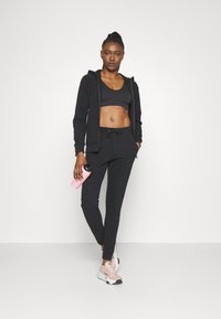 Fekete sportos kapucnis felső zsinórral, hozzá illő fekete crop top és melegítőnadrág. Rózsaszín vizes palack a jobb kézben. Világos rózsaszín sportcipők.