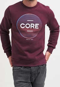 Sudadera burdeos con un diseño gráfico redondo que presenta líneas concéntricas y la palabra "CORE" en blanco. Textura suave, estilo cuello redondo.