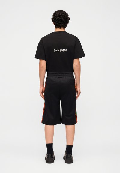 Άνδρας όρθιος με την πλάτη γυρισμένη, φοράει μαύρο μπλουζάκι Palm Angels, μαύρα σορτς με χρωματιστές ρίγες, μαύρες κάλτσες και μαύρα παπούτσια σε λευκό φόντο.