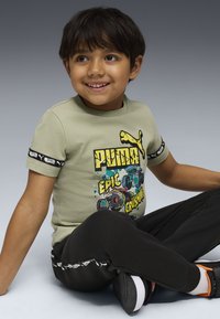 Lächelnder Junge, der im Schneidersitz sitzt, tr�ägt ein hellgrünes Puma-Graphic-T-Shirt mit Monstertrucks und schwarze Hose mit Puma-Logos.