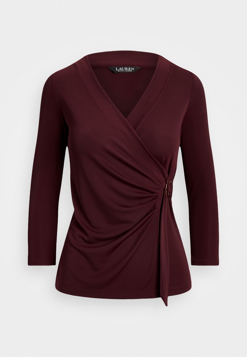 Haut long à manches burgundy enroulé avec un décolleté en V, présentant un détail torsadé sur le côté et un accent de quincaillerie décoratif à la taille. Texture de tissu lisse.