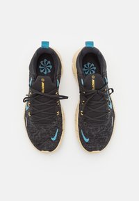 Une paire de chaussures de course Nike noires et beiges avec des logos teal, des lacets noirs et un design floral sur les semelles intérieures, vue de dessus.