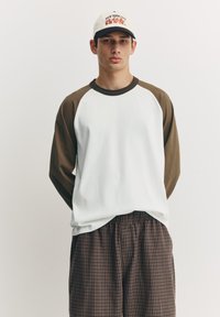 Cămașă albă cu mâneci lungi maro raglan, asortată cu pantaloni scurți maro cu carouri. Șapcă inscripționată cu "NEW YORK CITY" în roșu și negru.