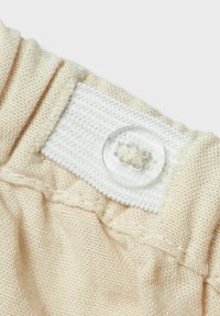 Lichtbeige stof met een gestructureerde weving, voorzien van een wit textielpatch en een transparante plastic knop met bloemdetail aan de rechterkant.