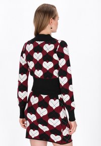Pletené šaty sveter v burgundskej farbe, zdobené srdcovým vzorom v čiernej a bielej. Má kontrastný čierny pás a rebrované manžety.