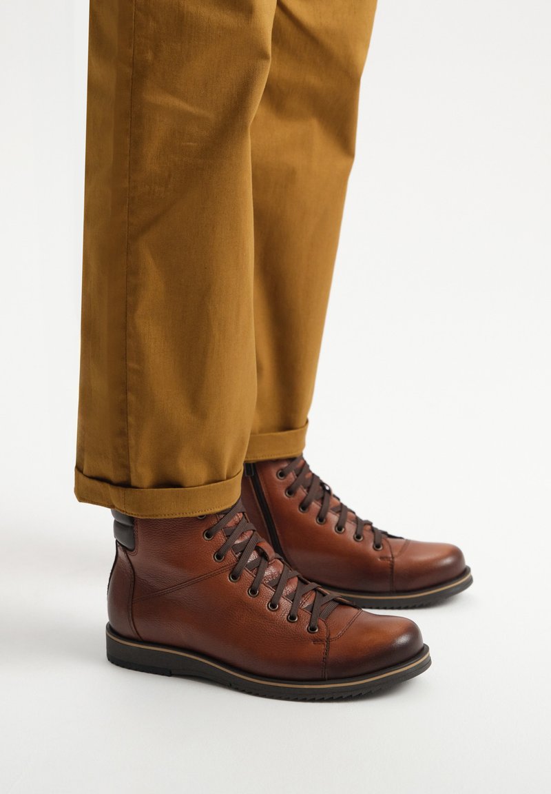 Brązowe skórzane botki za kostkę z okrągłym noskiem, gumową podeszwą i sznurowanym zapięciem. Nosi się je z podwiniętymi beżowymi spodniami, które podkreślają swobodny styl.