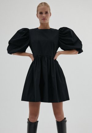 Kobieta stoi z rękami na biodrach, ubrana w krótką czarną sukienkę z bufkami i czarne kozaki do kolan na tle jednolitym.