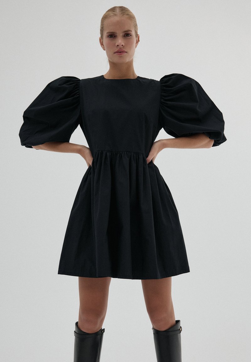 Kobieta stoi z rękami na biodrach, ubrana w krótką czarną sukienkę z bufkami i czarne kozaki do kolan na tle jednolitym.