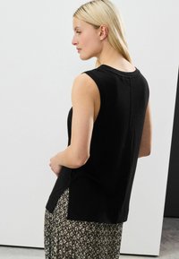 Top nero senza maniche con scollo rotondo, fessure laterali e una vestibilità comoda, realizzato in un tessuto morbido. Il modello lo indossa con una gonna lunga fantasia.