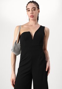 Sorte jumpsuit med én skulder og dyp V-hals, tettsittende overdel, bred bein design, sammen med en metallisk sølv håndveske.