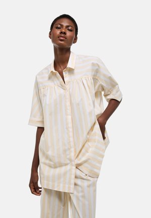 Femme portant une chemise ample à rayures verticales beige et blanches et un pantalon large assorti, debout avec une main dans la poche.