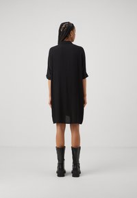 Schwarzes Kurzarmkleid mit lockerem Schnitt, runden Ausschnitt und leicht geschwungenem Saum. Getragen mit hohen schwarzen Stiefeln.